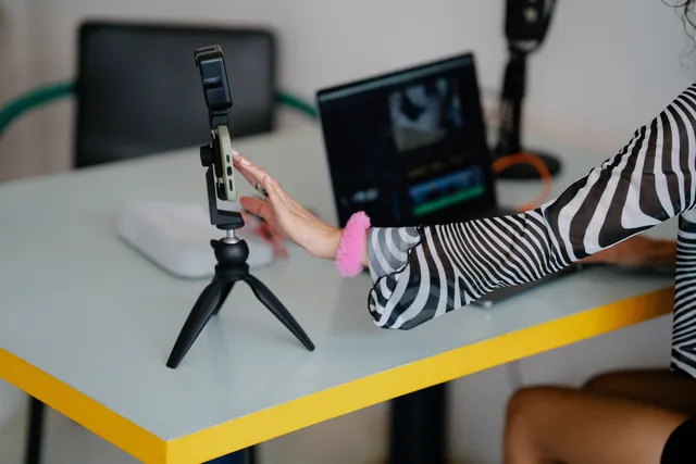Woman adjusting smartphone on tripod with laptop setup for video editing. Influencer agency tool implementation: Traackr access and release checks